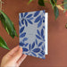 Hand holding a blue floral-patterned card against a brown background with green leaves.