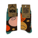 A pair of patterned crew socks shown front and back with attached kraft paper tags. The socks have a dark brown base with large abstract smiley-face shapes in pink, orange, yellow, and teal, plus teal cuff sections.