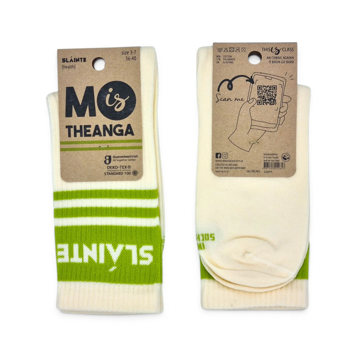 Two pairs of cream-colored crew socks with green stripes and white text 'SLAINTE', displayed on their original kraft paper packaging against a white background. The brand 'MO is THEANGA' is clearly visible on the left package.