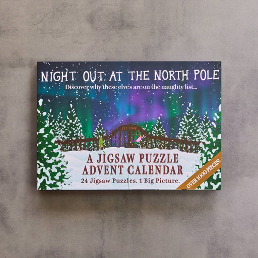 A jigsaw puzzle advent calendar titled 'Night Out at the North Pole' resting on a textured gray surface. The box features a colorful winter night scene with aurora lights, a bridge, and snowy trees, with text indicating 24 jigsaw puzzles in 1 big picture.