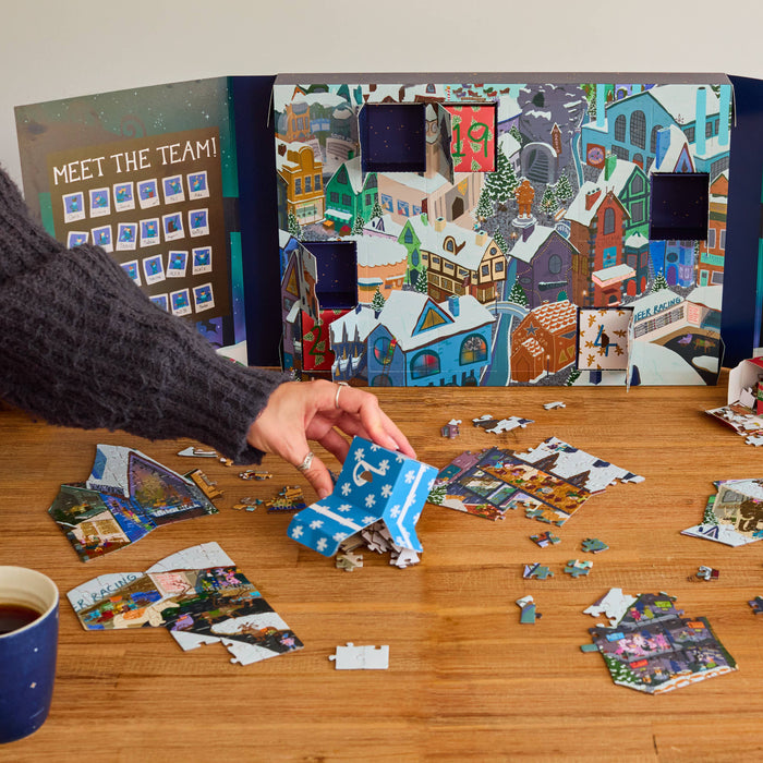 A person's hand is assembling a jigsaw puzzle from a blue and white patterned pouch on a wooden table. Behind the hand is a large illustrated Advent calendar panel depicting a snowy Christmas village, with scattered puzzle pieces around it, a coffee mug, and a bowl of nuts.
