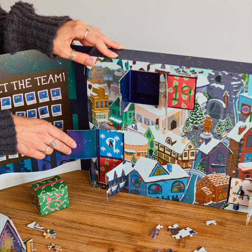 A person's hands are shown assembling a complex jigsaw puzzle featuring a colorful winter village scene with houses, trees, and snowy roofs. Several completed puzzle pieces are scattered on a wooden table next to a wrapped gift box.
