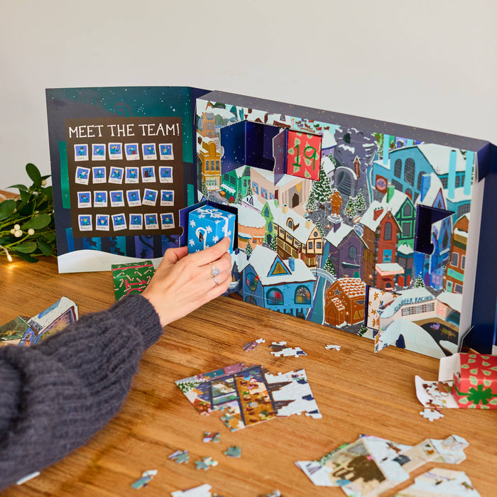 A person's hand opens a colourful Christmas advent calendar on a wooden table, with jigsaw puzzle pieces scattered around and a bowl of nuts nearby.