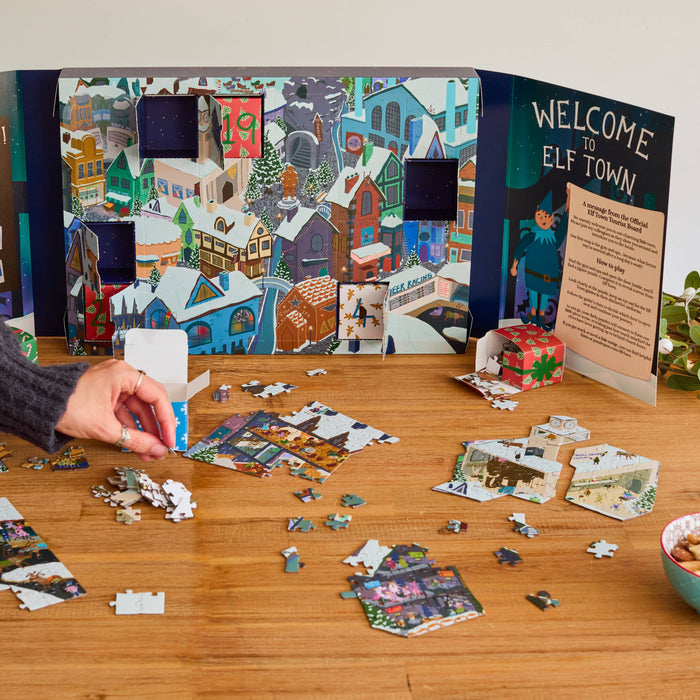 A person's hands are shown assembling a jigsaw puzzle on a wooden table, with an illustrated Advent calendar displaying 'Meet the Team' and 'Welcome to Elf Town' behind it. The scene includes a blue mug of coffee and a bowl of nuts on the table.