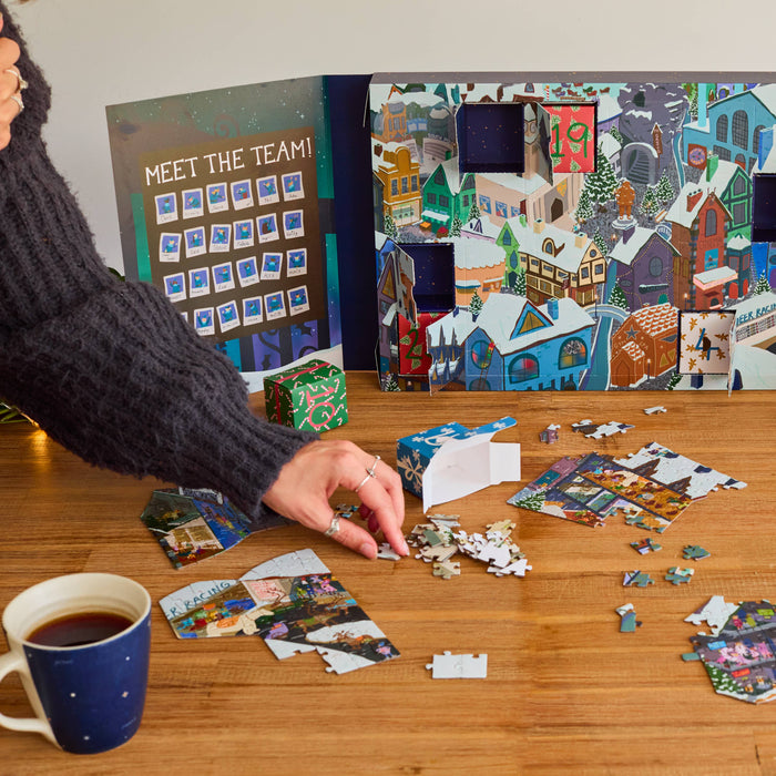 A person's hand is assembling a colorful jigsaw puzzle on a wooden table. The puzzle features an illustrated winter village scene with snow-covered houses, trees, and a festive town sign reading 'Welcome to Elf Town.