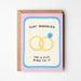 A pink greeting card with a kraft envelope, featuring a cartoon illustration of two interlocking yellow wedding rings with a small blue gemstone and the text 'JUST MARRIED HAS A NICE RING TO IT'.