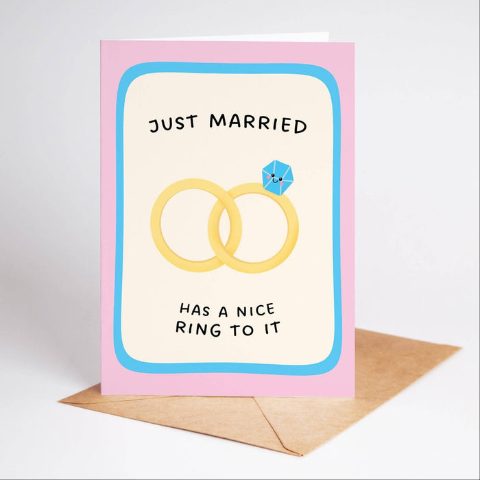 A pink greeting card with a light blue border and black text reading 'JUST MARRIED HAS A NICE RING TO IT,' featuring an illustration of two yellow wedding rings with a small blue gemstone. A brown envelope sits behind the card on a light gray background.
