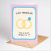 A pink greeting card with a light blue border and black text reading 'JUST MARRIED HAS A NICE RING TO IT,' featuring an illustration of two yellow wedding rings with a small blue gemstone. A brown envelope sits behind the card on a light gray background.