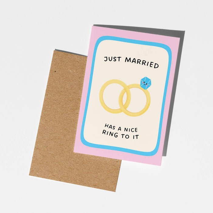 A square greeting card with a kraft envelope, featuring the text 'JUST MARRIED HAS A NICE RING TO IT' and an illustration of two interlocking wedding rings with a small blue gem, shown on a light gray background.