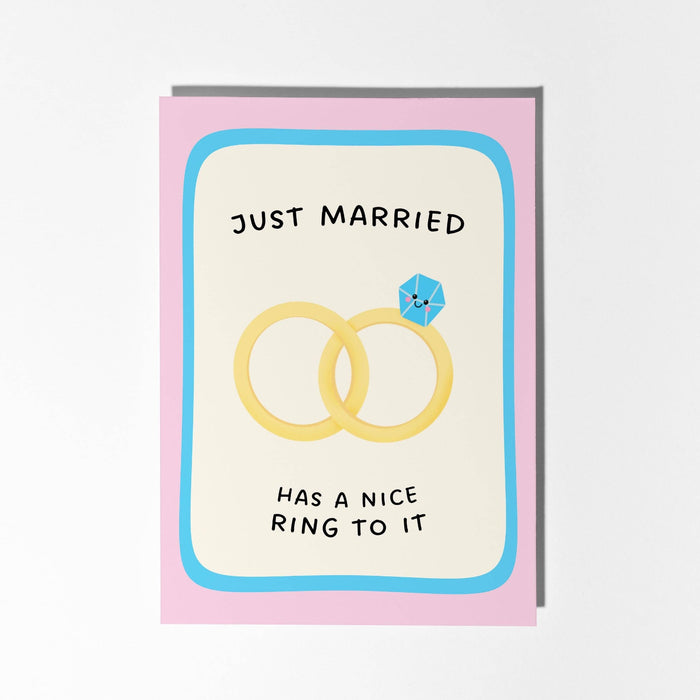 A pink greeting card with a blue border and a cream center panel that reads 'JUST MARRIED HAS A NICE RING TO IT,' featuring two interlocking yellow rings and a small blue gemstone illustration.