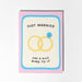 A pink greeting card with a blue border and a cream center panel that reads 'JUST MARRIED HAS A NICE RING TO IT,' featuring two interlocking yellow rings and a small blue gemstone illustration.
