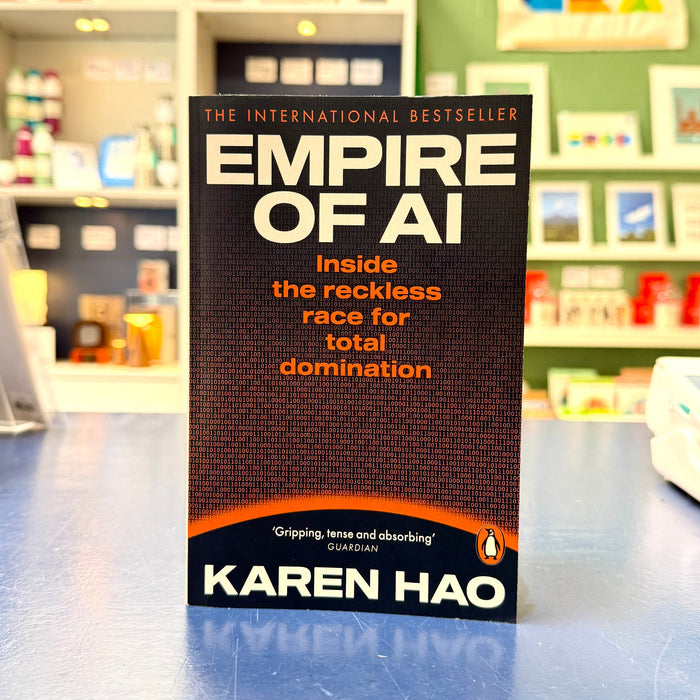 The book 'Empire of AI' by Karen Hao stands upright on a blue countertop in a retail store. The cover is predominantly black with white and orange text, and the Penguin logo is visible at the bottom.