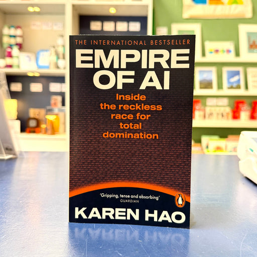 The book 'Empire of AI' by Karen Hao stands upright on a blue countertop in a retail store. The cover is predominantly black with white and orange text, and the Penguin logo is visible at the bottom.