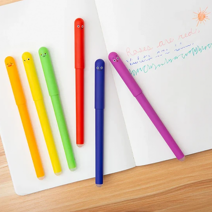 Six colorful novelty gel pens with cartoon faces are arranged on an open notebook on a wooden table. The pens are yellow, orange, green, red, blue, and purple, with the notebook showing some handwritten text.