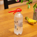 A clear plastic soda bottle with a bright red cap and red carrying loop, standing on a wooden table with a banana and flowers in the background.