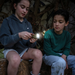 Two children sitting outdoors on a log at night, with one holding a small flashlight. One child wears a dark blue hoodie and white shorts, and the other wears a green long-sleeve shirt and light blue jeans.