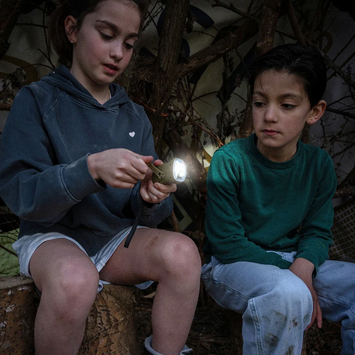 Two children sitting outdoors on a log at night, with one holding a small flashlight. One child wears a dark blue hoodie and white shorts, and the other wears a green long-sleeve shirt and light blue jeans.