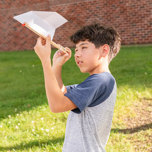 Child holding a homemade paper airplane outdoors on a grassy area with a brick wall in the background.