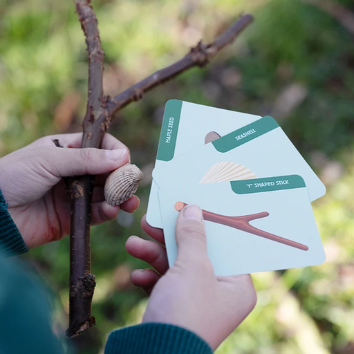 A person outdoors holds a twig branch in one hand and several illustrated cards in the other, including a card labeled 'MAKE A SEASHELL' and another labeled '7" SHAPED STICK'.