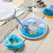 A circular embroidery hoop displays a completed bird design in blue, gray, white, and yellow thread, placed on a light wooden table with embroidery tools, yarn skeins, and a glass of water.