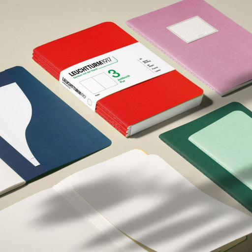 A set of Leuchtturm1917 pocket notebooks arranged on a light beige surface, including a red stack with a white paper band, a pink notebook, a navy notebook, a green notebook, and an open white notebook in the foreground.
