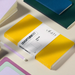 A Leuchtturm1917 notebook in a bright yellow cover with a white paper band label, styled on a light beige surface with other notebooks and a pastel pen around it.