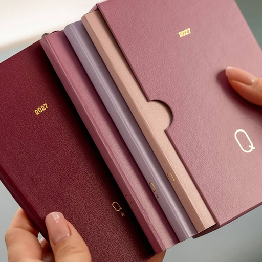 Four textured 2027 pocket calendars in shades of burgundy, mauve, dusty rose, and beige, held in hands to show their slim profiles and card-slot covers.