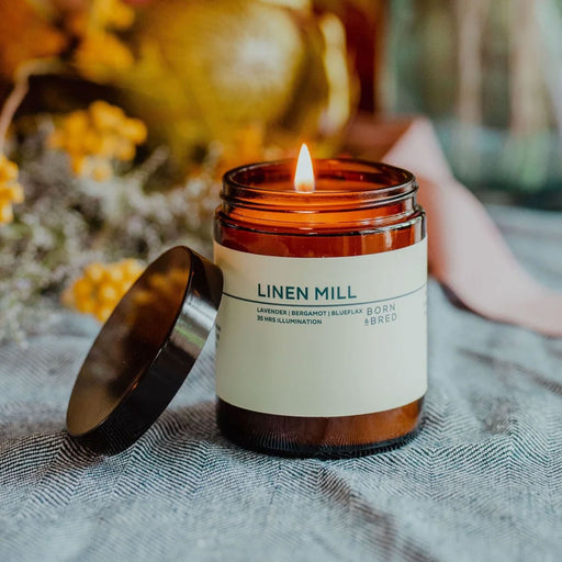 lit candle in brown glass jar with lid to left. jar has a cream label with green text