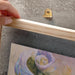 Hand holding a wooden frame with a metallic hook on a concrete surface with a painting of flowers.