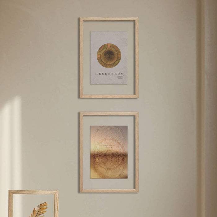 Two framed abstract art prints with gold-toned designs hang on a beige wall above a white table. One frame contains a feather illustration, and the other features circular geometric artwork.