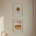 Two framed abstract art prints with gold-toned designs hang on a beige wall above a white table. One frame contains a feather illustration, and the other features circular geometric artwork.