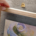 Hand holding a wooden frame with a metallic hook on a concrete surface with a painting of flowers.