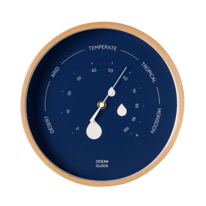 A round ocean clock with a light wood frame and a deep navy blue dial. The dial features white text and markings for temperature zones (Temperate, Tropical, Monsoon, Desert, Arid) and water droplet indicators, with white hands and a central gold accent.