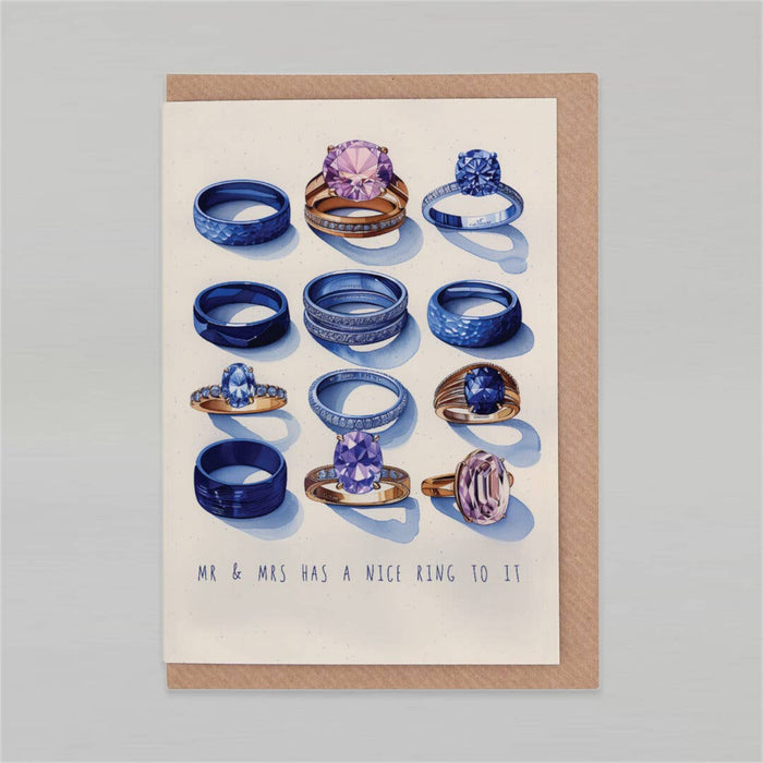 A square greeting card with a kraft paper border, featuring an illustration of various blue and purple gemstone rings on an off-white background, with the text 'MR & MRS HAS A NICE RING TO IT' printed at the bottom. The card is displayed flat against a neutral gray background.