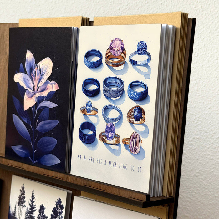 An open art book or journal displayed on a shelf, showing illustrated covers: one with a dark blue lily on black, and one with multiple illustrated rings in blue, purple, and gold on a cream background. The book is photographed against a light textured wall.