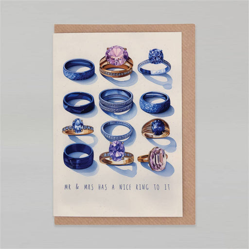 A square greeting card with a kraft paper border, featuring an illustration of various blue and purple gemstone rings on an off-white background, with the text 'MR & MRS HAS A NICE RING TO IT' printed at the bottom. The card is displayed flat against a neutral gray background.
