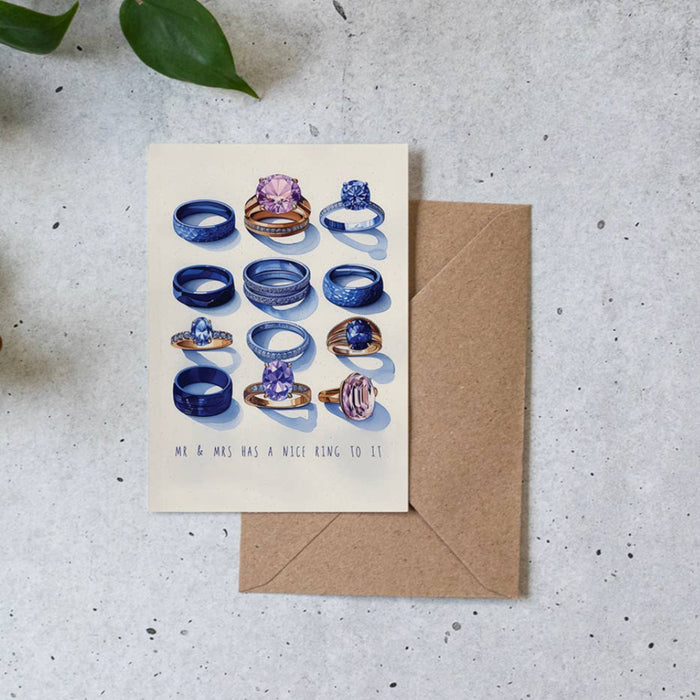 A square cream greeting card with an illustration of multiple blue and purple gemstone rings, placed on top of a brown kraft envelope on a light speckled surface with green leaves at the edge.