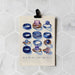 A printed card clipped at the top against a white hexagonal tile wall. The card shows illustrated blue and purple rings with the text 'MR & MRS HAS A NICE RING TO IT'.