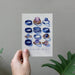 A hand holds a white art print featuring watercolor illustrations of various blue and purple gemstone rings, with the text 'A MRS HAS A NICE KING TO IT' at the bottom, set against a textured gray wall with green plant leaves in the foreground.