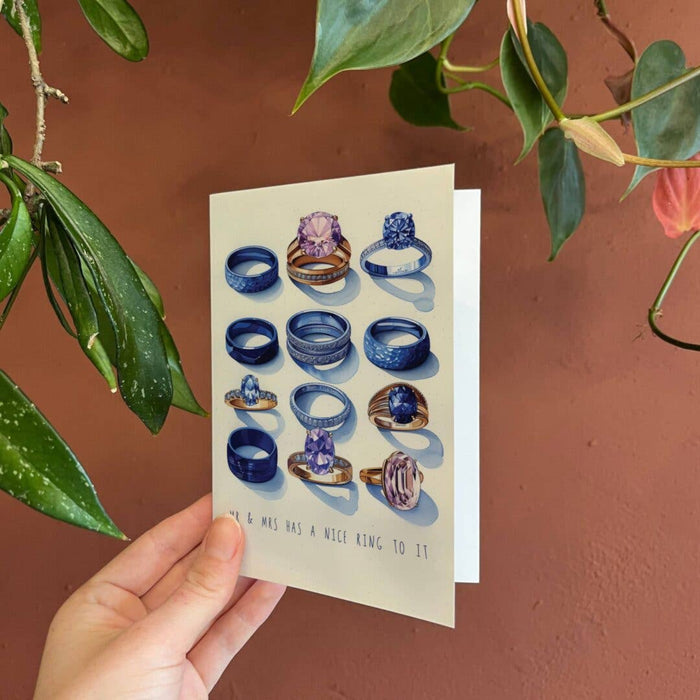 A hand holds a cream-colored greeting card featuring illustrations of various blue and purple gemstone rings, set against a terracotta wall with green plant leaves.