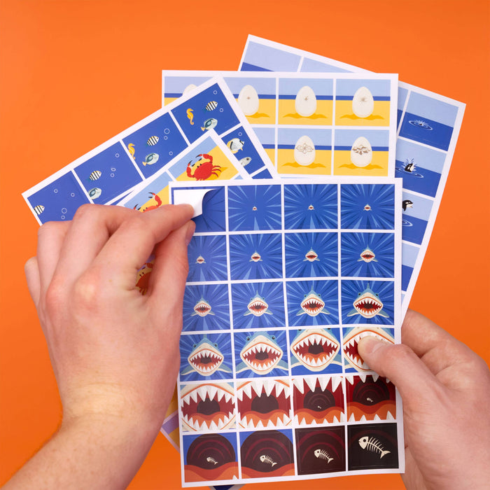 Two hands holding a fanned-out set of children's puzzle or activity sheets with shark and fish illustrations, against a solid orange background.