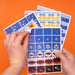 Two hands holding a fanned-out set of children's puzzle or activity sheets with shark and fish illustrations, against a solid orange background.