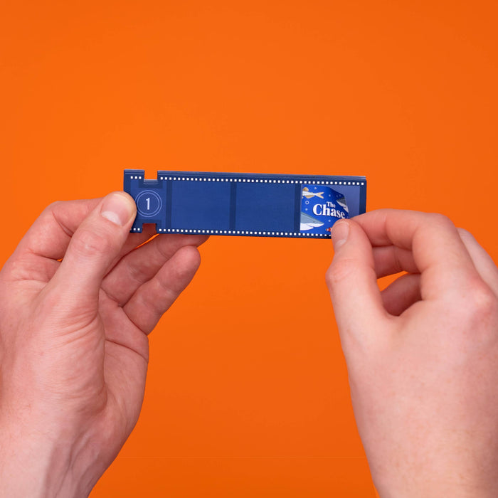 A person's hands holding a miniature blue film strip prop against a solid orange background. The prop features a 'The Chase' logo on the right side and a '1' on the left.