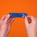 A person's hands holding a miniature blue film strip prop against a solid orange background. The prop features a 'The Chase' logo on the right side and a '1' on the left.