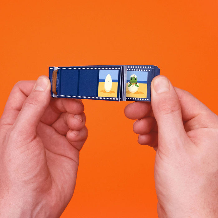 Two hands holding a miniature blue film camera made from a strip of old photo negatives, set against a solid orange background.