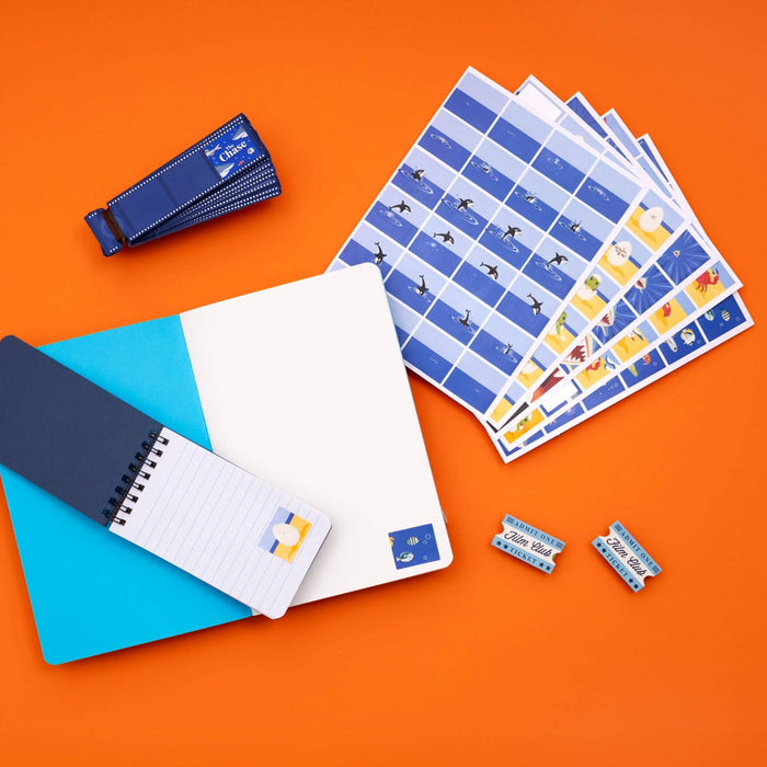 A flat-lay arrangement of blue-themed stationery and small accessories on a solid orange background, including a notebook, a spiral-bound notepad, sticker sheets, a wrist strap, and two small rectangular items.