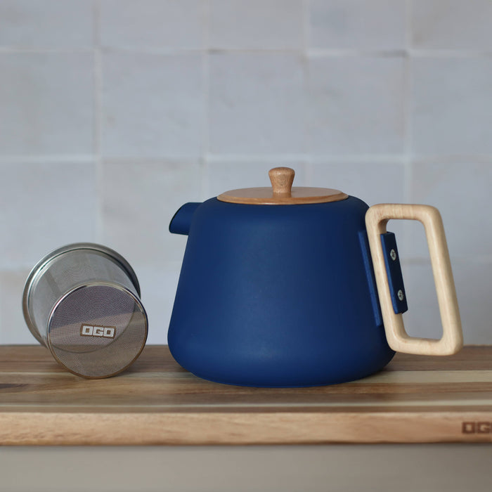 Blue teapot with wooden handle and lid on a wooden surface, with a white tiled wall in the background.