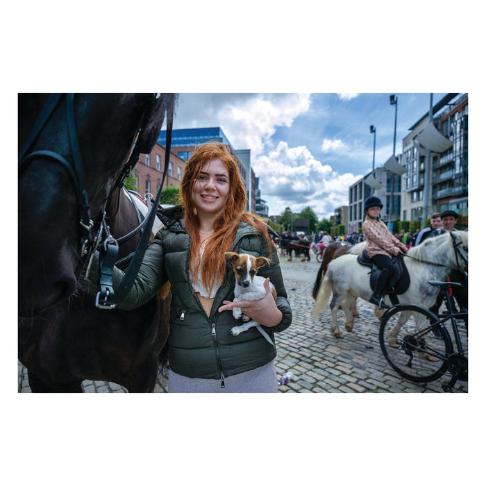 Woman with red hair holding a dog, standing next to a horse on a city street.