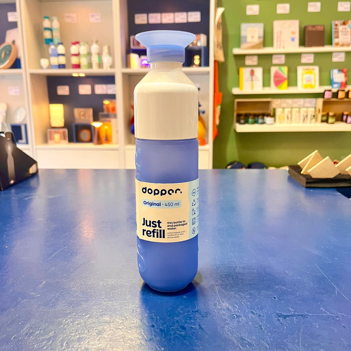 Blue Dopper water bottle with white cap on a blue surface, with shelves and products in the background.