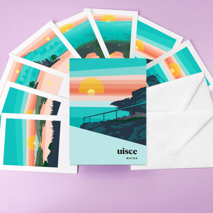 A set of illustrated postcards and white envelopes arranged on a lavender background. The postcards feature a stylized sunset landscape with teal water, pink and orange sky layers, and a yellow sun, with the brand name 'uisce WATER' printed on the front card.
